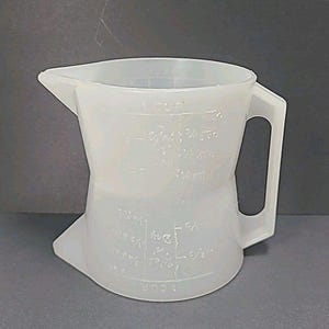 May include: A translucent white plastic measuring jug with a handle and a pouring spout. The jug has measurement markings in cups, fluid ounces, and fractions. The handle is rectangular and attached to the side of the jug.