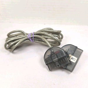 May include: A coiled silver-toned cable with a translucent gray plastic casing, featuring two distinct semi-circular connectors at one end. The cable is secured with a small purple rubber band, and the internal wiring is visible through the shell.