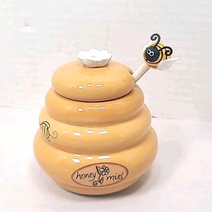May include: A ceramic honey pot shaped like a beehive, with a matching lid and a wooden honey dipper. The pot is yellow with a floral design and the words "honey miel". The dipper is topped with a cartoon bee.