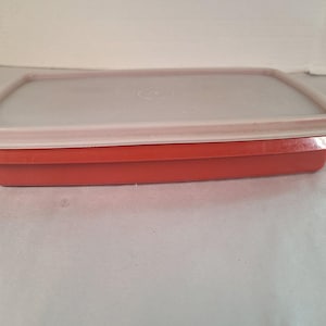 May include: A rectangular, red plastic container with a light gray lid. The lid has a white rim and a circular logo in the center. The container is designed for food storage.