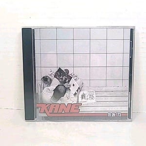 Puede incluir: Una caja de CD con el álbum "19 in '73" de Kane. La portada muestra a una persona leyendo una revista en un banco. El título del álbum y el nombre del artista están en texto rojo y blanco.