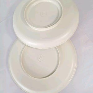 May include: Two off-white plastic plates are stacked, showcasing their underside. The plates have a circular design with a raised center and a slightly curved edge. The plates are a solid, neutral color.