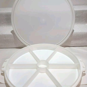 May include: A white plastic serving tray with a lid. The tray is divided into six sections and has a round shape. The lid is also round and white. The tray has handles on the sides.