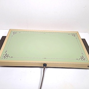 May include: Rectangular electric griddle with a light green surface and decorative black corner accents. The griddle has a light wood-colored frame and dark brown handles on each side. A brown power cord extends from the griddle.