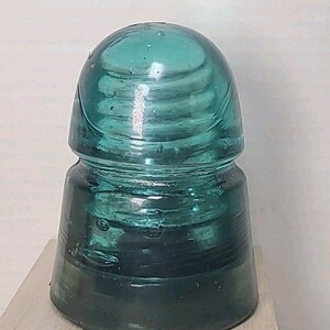 Puede incluir: Un aislador de vidrio de color verde azulado con una parte superior redondeada y una base cilíndrica. El vidrio tiene una apariencia texturizada y arremolinada, exhibido sobre una superficie de madera. Artículo vintage.