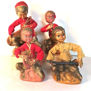 May include: Four vintage figurines depicting characters in red or tan long-sleeved tops and dark trousers. Each figure is seated on a textured base, with some holding musical instruments like a lyre or a flute, and others holding a sword.