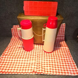 Könnte beinhalten: Ein Vintage-Picknick-Set mit zwei rot-weißen Isolierflaschen mit Griffen, einer roten Kunststoff-Lunchbox und einem braunen Kunstleder-Etui. Das Set ist auf einer rot-weiß karierten Picknickdecke angeordnet.