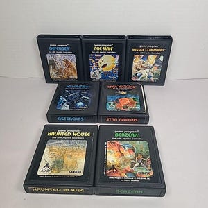 May include: A collection of seven Atari game cartridges, including "Defender," "Pac-Man," "Missile Command," "Asteroids," "Star Raiders," "Haunted House," and "Berzerk." Each black cartridge features colorful game art and text labels.