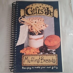 Op de afbeelding: Een spiraalgebonden kookboek met de titel "Gifts in a Jar: Muffins & Breads". De omslag toont een pot met gelaagde ingrediënten, een muffin en tekst. De pot is versierd met een sterrenpatroon stoffen deksel en raffia. De ondertitel van het boek luidt "Recepten om je eigen cadeaus te maken."