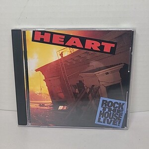 Op de afbeelding: Een cd-hoesje met de bandnaam "HEART" in rode letters. De albumhoes toont een beschadigd gebouw tegen een oranje en gele lucht. Een blauwe sticker leest "ROCK THE HOUSE LIVE!"