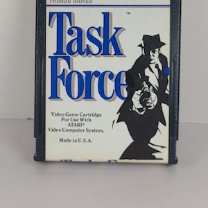 Puede incluir: Cartucho de videojuego Atari "Task Force" de Froggo Games. La etiqueta azul y blanca muestra el título del juego y una silueta de un hombre con sombrero sosteniendo un arma. Fabricado en EE. UU. para el sistema Atari Video Computer.