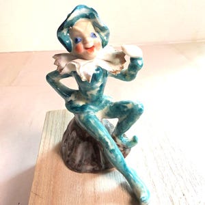 Può includere: Una statuetta in porcellana raffigurante un giullare con un costume maculato blu e bianco e un cappello abbinato. Il giullare ha un colletto bianco arricciato con bordo dorato, occhi blu, bocca rossa ed è seduto su una base marrone rocciosa.