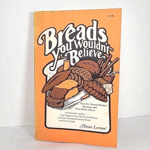 Peut inclure: Un livre de cuisine vintage intitulé "Breads You Wouldn't Believe" avec une couverture orange. La couverture présente des illustrations de pains variés et le texte "Easy-Do Natural Recipes". Le livre contient 119 recettes.