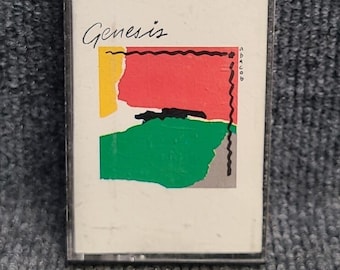 Può includere: Una custodia per cassette nera con un'etichetta bianca che presenta la parola "Genesis" in scrittura corsiva nera. L'etichetta presenta anche un design astratto con forme gialle, rosse, verdi, nere e grigie.