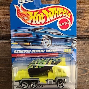 May include: A Hot Wheels Oshkosh Cement Mixer toy car in its original packaging. The cement mixer is yellow and black, with silver wheels. The packaging is blue with the Hot Wheels logo and text that reads "Guaranteed for Life."