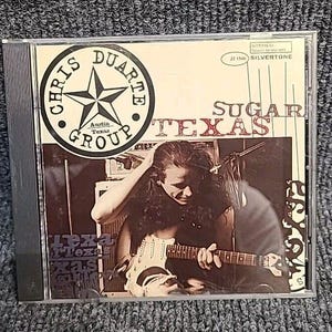 May include: A CD case with a black and white photo of a man playing a white electric guitar. The CD case has a black and white logo that says "Chris Duarte Group" and the text "Sugar Texas".