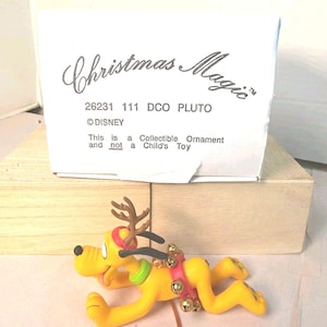 Pode incluir: Uma figura decorativa amarela do Pluto com chifres de rena castanhos, uma coleira verde e um arnês vermelho com pequenos sinos dourados. A caixa branca atrás diz: Christmas Magic, 26231 111 DCO PLUTO, DISNEY, This is a Collectible Ornament and not a Child's Toy.