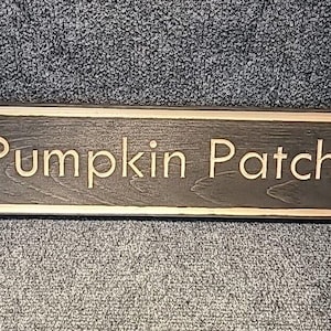 Op de afbeelding: Een rechthoekig houten bord met de woorden "Pumpkin Patch" in een lichte kleur gesneden. Het bord heeft een donkerbruine achtergrond en een lichtgekleurde rand. Het bord ligt op een gestructureerd grijs oppervlak.