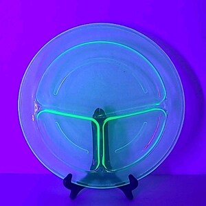 May include: A clear, divided glass plate with a three-section design. The plate has a bright green glow, with a central support structure. The plate is displayed on a black stand against a purple background.