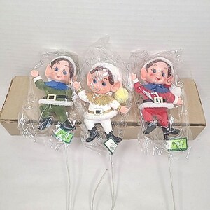 Könnte beinhalten: Drei Vintage-Weihnachtselfen-Figuren in Kunststoffverpackung. Jeder Elf hat ein anderes Outfit: grün, weiß und rot. Sie haben rosige Wangen, blaue Augen und weiße Hüte. Die Figuren sind an dünnen Metalldrähten befestigt.