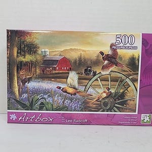May include: A 500-piece jigsaw puzzle featuring a rural landscape with a red barn, deer, and pheasants. The puzzle box has the "Artbox" logo and the artist's name, Lee Radcliff. The puzzle is titled "Coming Home".