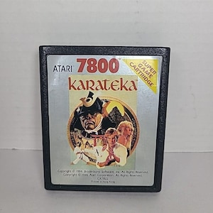 Może przedstawiać: Czarny kartridż do gry wideo Atari 7800 Karateka. Etykieta zawiera okrągłą ilustrację mężczyzny w kapeluszu, dwóch osób i tytuł gry. Kartridż jest oznaczony jako "Super Game Cartridge".