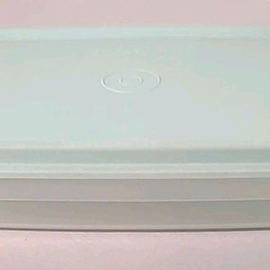 Puede incluir: Un recipiente rectangular de plástico azul claro con una tapa a juego. La tapa tiene una parte superior ligeramente redondeada y un emblema circular en el centro. El recipiente tiene un diseño acanalado en los lados.