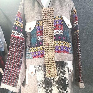 May include: A gray jacket with colorful patchwork details. The jacket features white pockets with brown buttons, and the sleeves have intricate woven patterns in red, black, and white. The front has a woven panel with brown buttons.