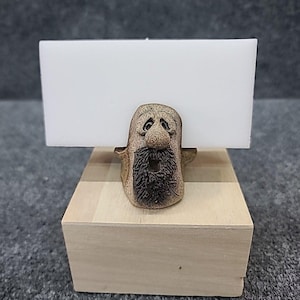 May include: A whimsical ceramic business card holder featuring a bearded face with a neutral color palette. The holder is mounted on a wooden block base, with a white card inserted. The design is detailed and handcrafted.