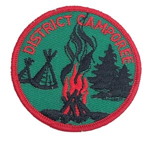 Może przedstawiać: Okrągła naszywka z zielonym tłem i czerwoną obwódką. Naszywka przedstawia ognisko z czerwonymi płomieniami, tipi i sosnami. Napis "DISTRICT CAMPOREE" jest wyhaftowany na górze na czerwono.