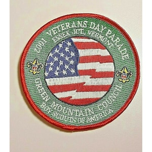 Könnte beinhalten: Runder Aufnäher mit rotem Rand und grünem Hintergrund. Der Aufnäher zeigt die amerikanische Flagge in der Mitte mit roten und weißen Streifen und einem blauen Sternenfeld. Der Text lautet "2001 Veterans Day Parade Essex Jct, Vermont" und "Green Mountain Council Boy Scouts of America."