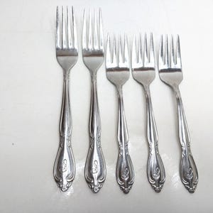 May include: A set of five silver-toned metal forks arranged in a row. Each fork features a polished finish, four tines, and a decorative handle with an embossed floral motif and a script letter E engraved at the base of the handle.