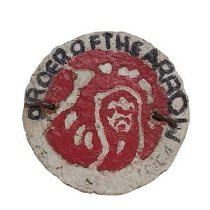 May include: A round, white, paper badge with a red, hand-painted design and the text "Brother of the Arrow" in black.