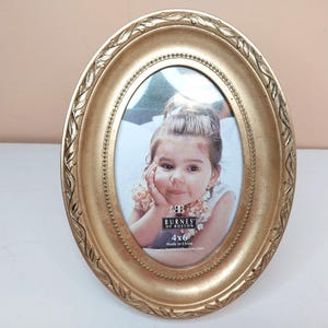 May include: An oval-shaped gold picture frame with a decorative leaf pattern along the outer edge and a beaded inner border. The frame holds a 4x6 inch photograph of a child with a hand resting on their chin, featuring the text Burnes of Boston.