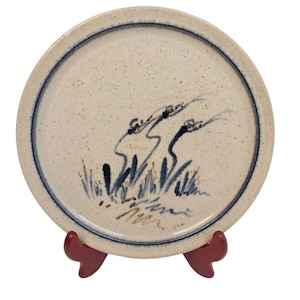 May include: A round, off-white ceramic plate with a blue rim and a hand-painted design of stylized plants. The plate is displayed on a red stand. The design features blue and brown brushstrokes.