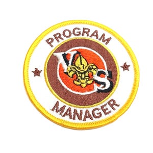 May include: A yellow and brown embroidered patch with the text "Program Manager" and a scout symbol in the center.