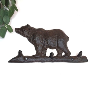 May include: A dark brown cast iron wall hook featuring a bear standing on a textured log base. The base includes three upward-curving hooks and two circular mounting holes for installation, with a detailed relief of a bear's fur and facial features.
