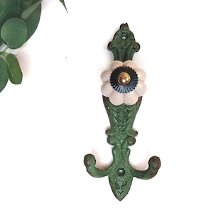 May include: A vertical wall hook featuring a green cast metal base with ornate scrollwork and two curved hooks at the bottom. A round, cream-colored ceramic knob with a dark brown center and a metallic gold-toned accent sits in the middle.
