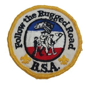 May include: A white and yellow embroidered patch with a red, white, and blue flag and the text "Follow the Rugged Road" and "RSA".