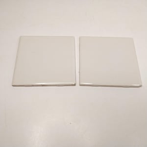 May include: Two square, off-white ceramic tiles with a smooth, glossy finish. Each tile features a flat surface and straight edges, presenting a clean, uniform appearance. The tiles are positioned side-by-side, showcasing their reflective, light-colored glaze.