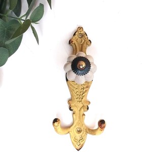 May include: A wall-mounted hook featuring a distressed yellow metal backplate with ornate scrollwork and a central, round, fluted ceramic knob in a pale gray finish with a small black and metallic center detail for hanging items.