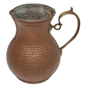 May include: A hammered copper pitcher with a brass handle. The pitcher has a bulbous body and a flared rim. The handle is curved and ornate. The copper has a warm, reddish-brown color. The pitcher is likely vintage or antique.