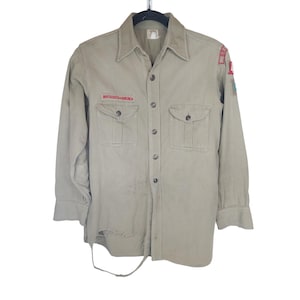 May include: A khaki button-down shirt with two chest pockets and patches. The shirt features the words "BOY SCOUTS OF AMERICA" in red on a patch. The shirt is suspended from a black hanger.