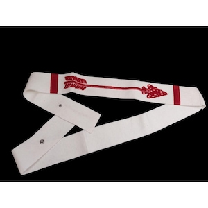 Puede incluir: Un cinturón de tela blanca con un diseño de flecha roja. La flecha está bordada en rojo y recorre toda la longitud del cinturón. El cinturón tiene dos cierres de botón metálicos.