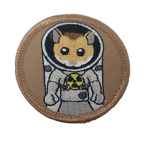 May include: A round, embroidered patch featuring a hamster in a spacesuit. The hamster has brown and tan fur, large black eyes, and is wearing a white spacesuit with a yellow hazard symbol. The patch has a tan border.