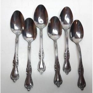 Puede incluir: Un juego de seis cucharas de metal plateado dispuestas en dos filas. Cada cuchara presenta un cuenco ovalado pulido y un mango decorado con un patrón floral en relieve que se extiende desde el cuello hasta la base redondeada del mango.