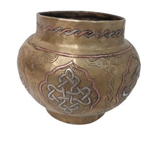 May include: A brass-colored, round vase with intricate etched designs. The vase features a decorative pattern of silver and copper accents. The design includes a central knot-like motif and a braided border near the rim. The vase has a wide body and a slightly flared opening.