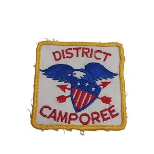Może przedstawiać: Kwadratowa naszywka z białym tłem i żółtą obwódką. Naszywka przedstawia niebieskiego orła z czerwoną i białą tarczą oraz dwiema czerwonymi strzałami. Napis "DISTRICT CAMPOREE" jest w kolorze czerwonym.