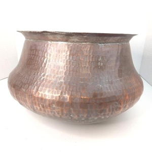 May include: A large, round, hammered copper pot with a wide, flared rim. The exterior surface features a distinct textured pattern created by manual hammering, showcasing the natural reddish-brown metallic finish of the copper material.