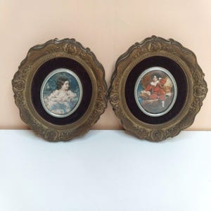 May include: Two circular wall frames with ornate, scalloped bronze-toned edges and dark velvet backing. Each frame contains an oval portrait of a child, one wearing white clothing and the other wearing a red outfit, set behind a silver-toned border.